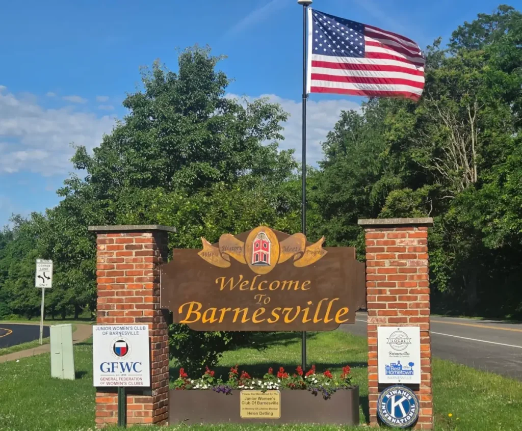Welcome to Barnesville sign, with an US flag on a pole behind it. Trees are in the distant background.